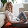 young woman sits on carpeted floor with her open laptop as she leans against her couch